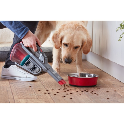 BLACKDECKER BHHV315B handstofzuiger zuigt dierenvoer op naast een rode kom op houten vloer, zijaanzicht.