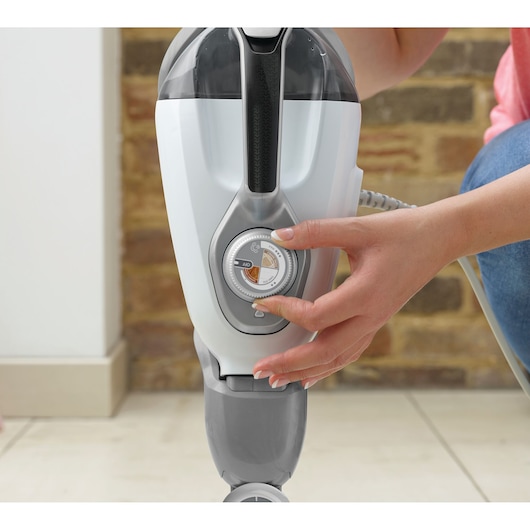 Close-up van een persoon die een regelknop op een BLACK+DECKER schoonmaakapparaat aanpast, met een bakstenen muur en een tegelvloer op de achtergrond.