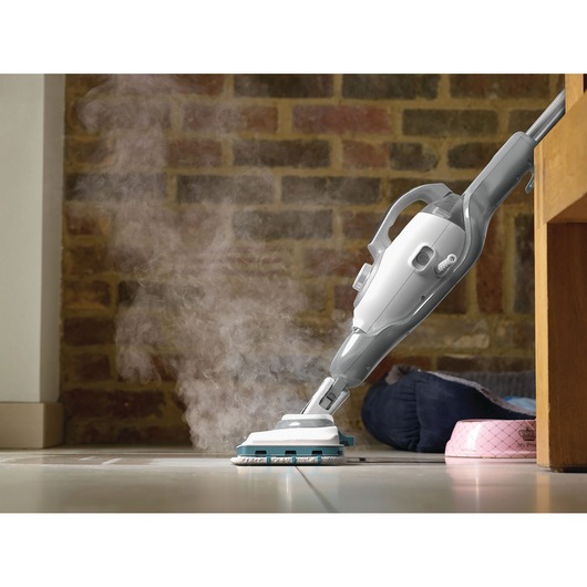 Een stoommop maakt een tegelvloer schoon met zichtbare stoom, naast een bakstenen muur en wat huisdieraccessoires.
