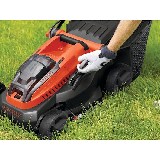 BLACKDECKER CLM3825L2 tondeuse gros plan, design orange et noir, main gantée réglant la hauteur sur l’herbe.