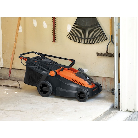 BLACK+DECKER CLM3825L2 tondeuse à gazon noir et orange, pliée pour rangement dans un garage à côté d’outils de jardin.