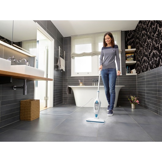 BLACKDECKER FSM13E1 balai vapeur nettoyant un sol en carrelage sombre de salle de bain, personne utilisant le produit près d'une baignoire.
