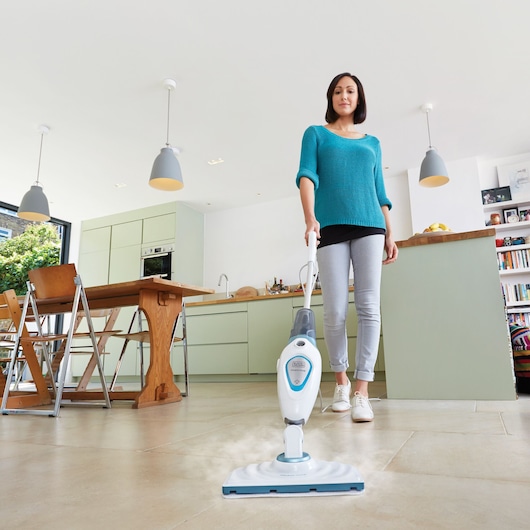 Une personne au visage flouté utilise un balai vapeur BLACK+DECKER FSM1605 pour nettoyer le sol carrelé dans une cuisine moderne. Le balai émet de la vapeur lorsqu'il est passé sur le sol.