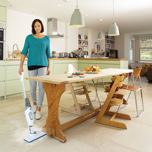 Une personne utilise un balai vapeur BLACK+DECKER FSM1605 pour nettoyer le sol dans une cuisine et salle à manger moderne. Il y a une table à manger en bois avec des fruits et des jouets dessus, entourée de plusieurs chaises. La cuisine est équipée de placards verts clair, d'appareils modernes et de lampes suspendues.