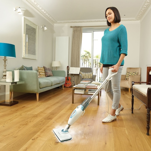 Une personne utilise un balai vapeur BLACK+DECKER FSM1605 pour nettoyer un sol en bois dans un salon moderne. La pièce est lumineuse et comprend un canapé, une guitare, une table avec des livres et de grandes fenêtres avec des rideaux.