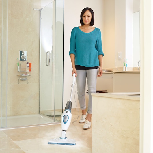 Une personne nettoie le sol de la salle de bain avec un balai vapeur BLACK+DECKER. La pièce comprend une douche, un lavabo et des surfaces carrelées beiges.