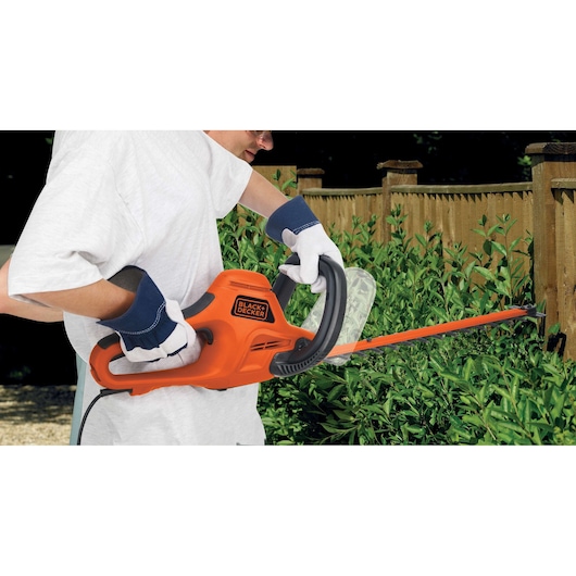 Foto van BLACK+DECKER GT7030 heggenschaar in gebruik, oranje elektrisch gereedschap snoeit groene haag bij houten schutting.