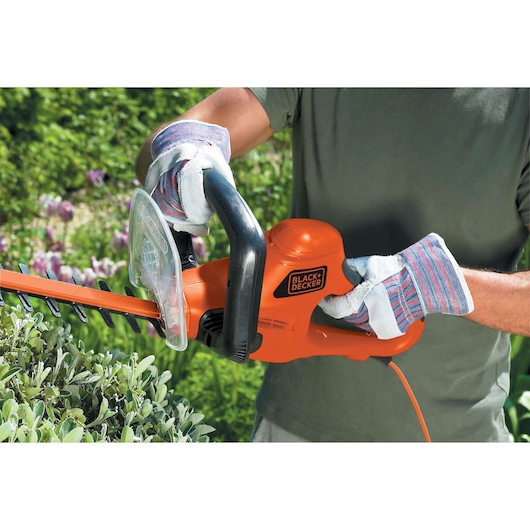 BLACK+DECKER GT7030 elektrische heggenschaar in gebruik, oranje behuizing, gebruiker met handschoenen, snoeit groene struiken buiten.