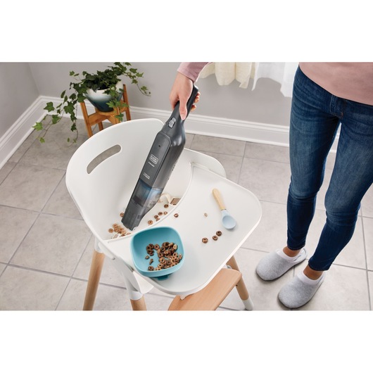 Aspirateur à main BLACKDECKER HLVC320J11 nettoyant des céréales sur le plateau d'une chaise haute dans une cuisine.