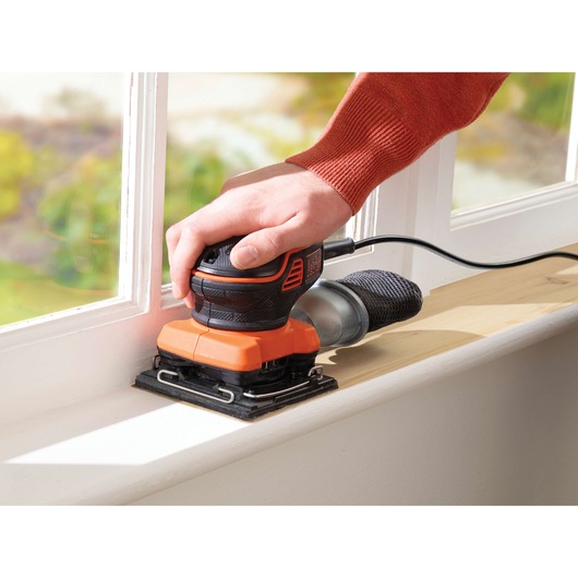 Foto van BLACK+DECKER KA450 vlakschuurmachine in gebruik op een houten vensterbank, met ergonomische greep en stofzak.