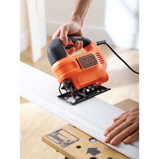 Foto van BLACK+DECKER KS701EK decoupeerzaag die wit hout zaagt, zwart-oranje gereedschap dat met de hand wordt gebruikt, binnen.