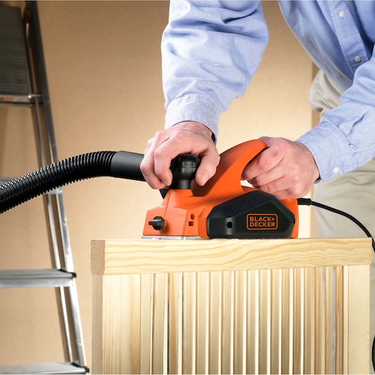 Rabot électrique BLACK+DECKER KW712 utilisé sur du bois, avec raccord pour aspirateur, vue rapprochée de côté.