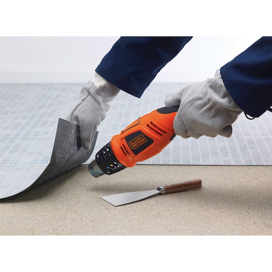 BLACKDECKER KX1693 oranje heteluchtpistool bij vloer verwijderen, met handschoenen en metalen schraper zichtbaar.