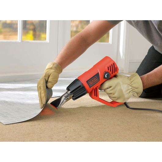 BLACKDECKER KX2001K heteluchtpistool gebruikt voor vloer verwijderen, oranje behuizing, hand met handschoen, in gebruik.