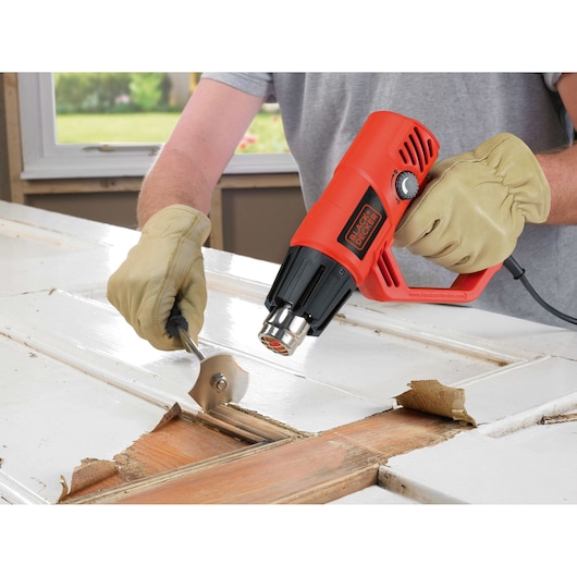 BLACKDECKER KX2001K heteluchtpistool gebruikt om verf van houten deur te verwijderen, close-up met handschoenen.