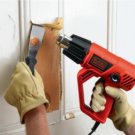 Foto van BLACKDECKER KX2001K heteluchtpistool gebruikt voor verfverwijdering op houten deur, vastgehouden door handschoenen.
