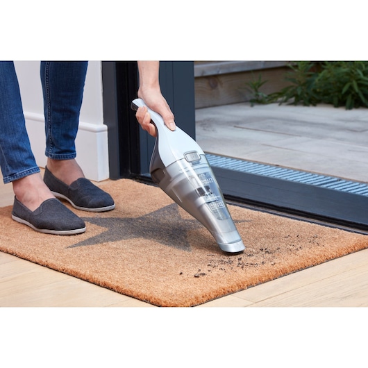 BLACKDECKER NVC215W handstofzuiger verwijdert vuil van een bruine deurmat bij een deuropening.