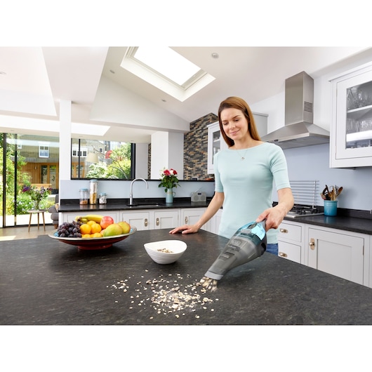 BLACKDECKER NVC220WBC handstofzuiger reinigt keukentafel met gemorste havermout naast fruitschaal.