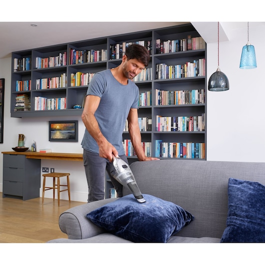 BLACKDECKER NVC220WC handstofzuiger gebruikt op een grijze bank met blauwe kussens in een moderne woonkamer.