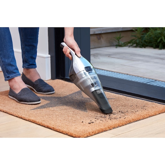 BLACK+DECKER NVC220WC handstofzuiger verwijdert vuil van een bruine deurmat bij een deuropening.