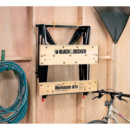 BLACKDECKER WM825 établi pliant fixé sur un mur en bois de garage, entouré d'un tuyau, d'un râteau et d'un vélo.